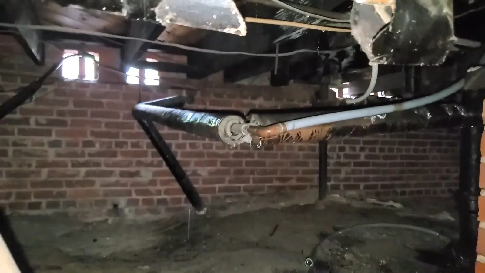 Black drain pipe and wrapped lines hanging under floor framing above a brick foundation wall