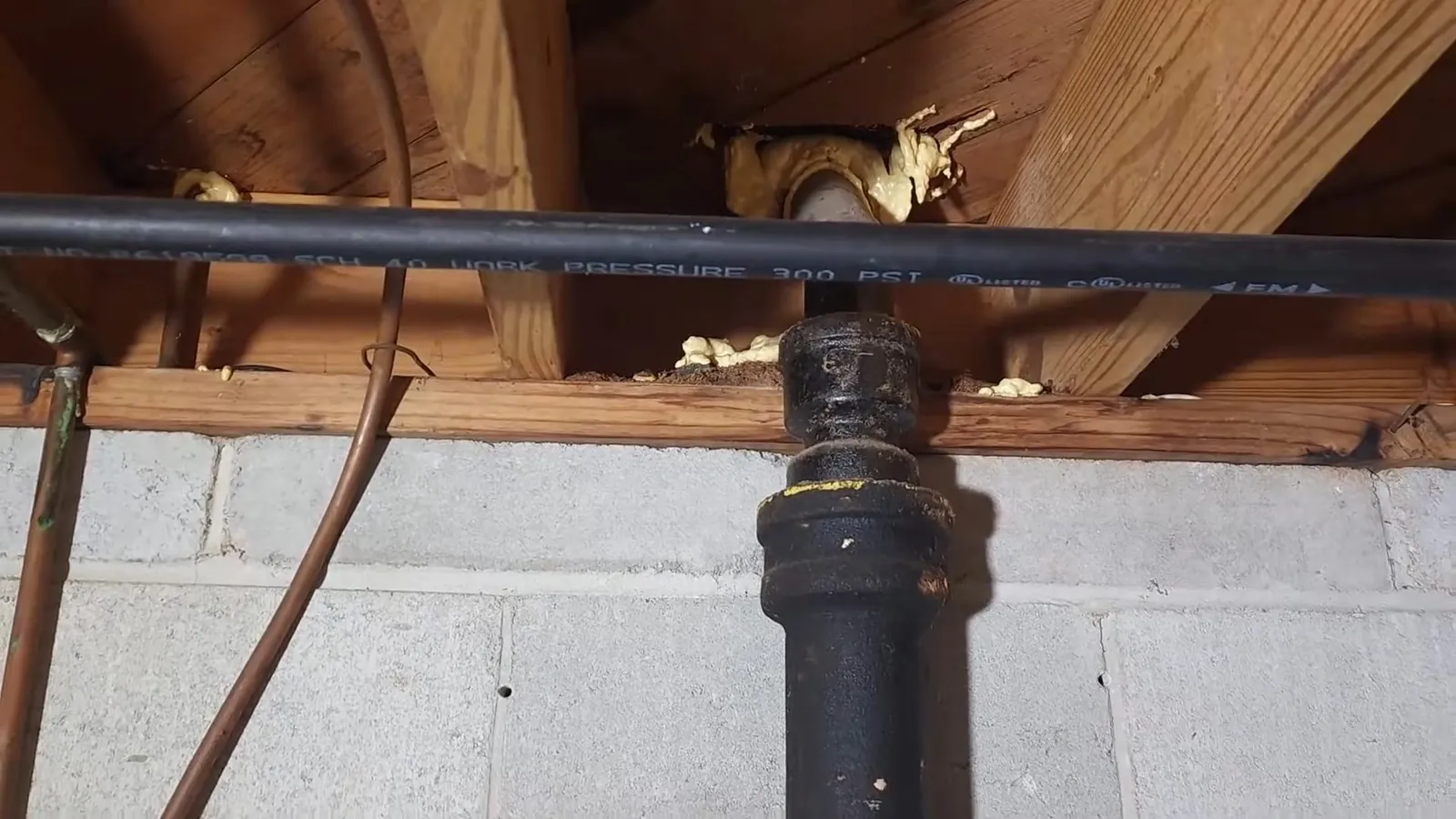 Black cast iron drain pipe with a brass fitting running under wooden floor joists above a block wall