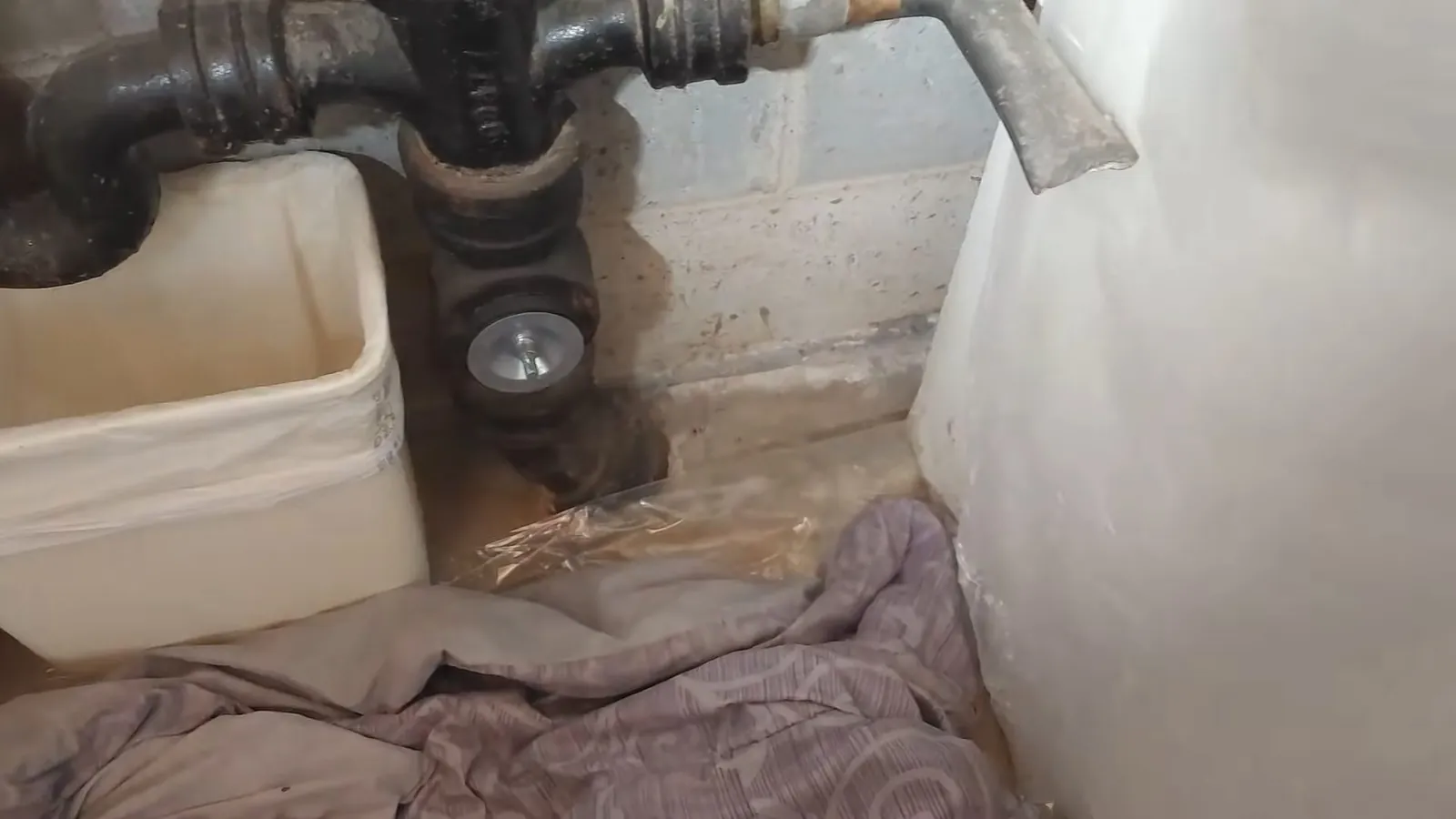 Black cast iron drain fittings above a white bucket on a wet basement floor, with a cloth lying under them
