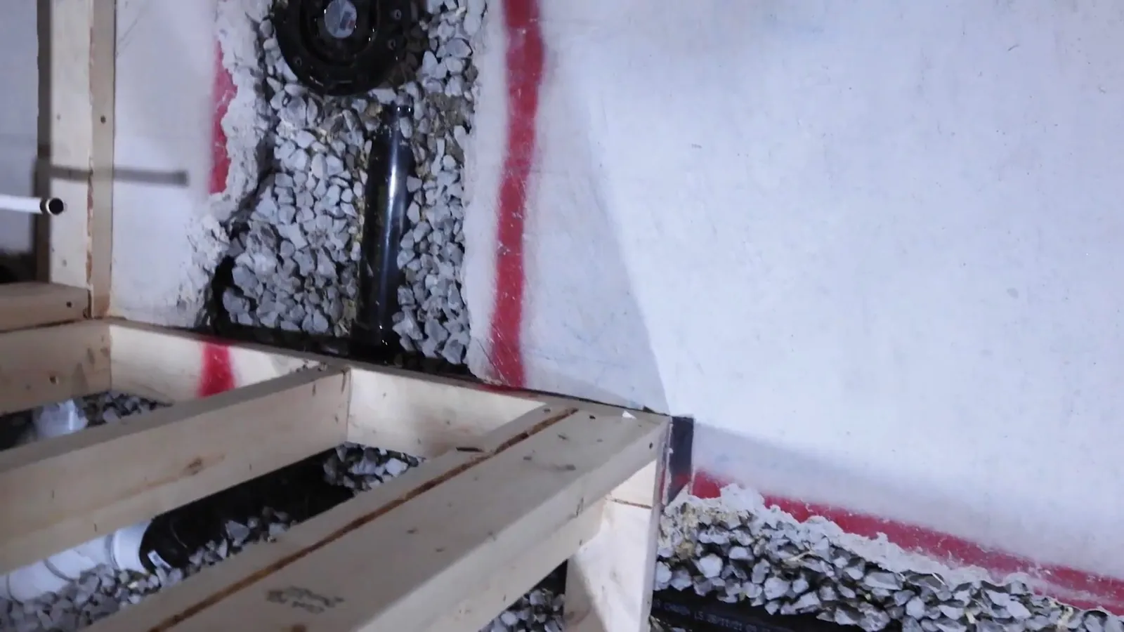 Black drain pipe set in gravel inside an opening cut into a concrete slab at the foot of a framed wall