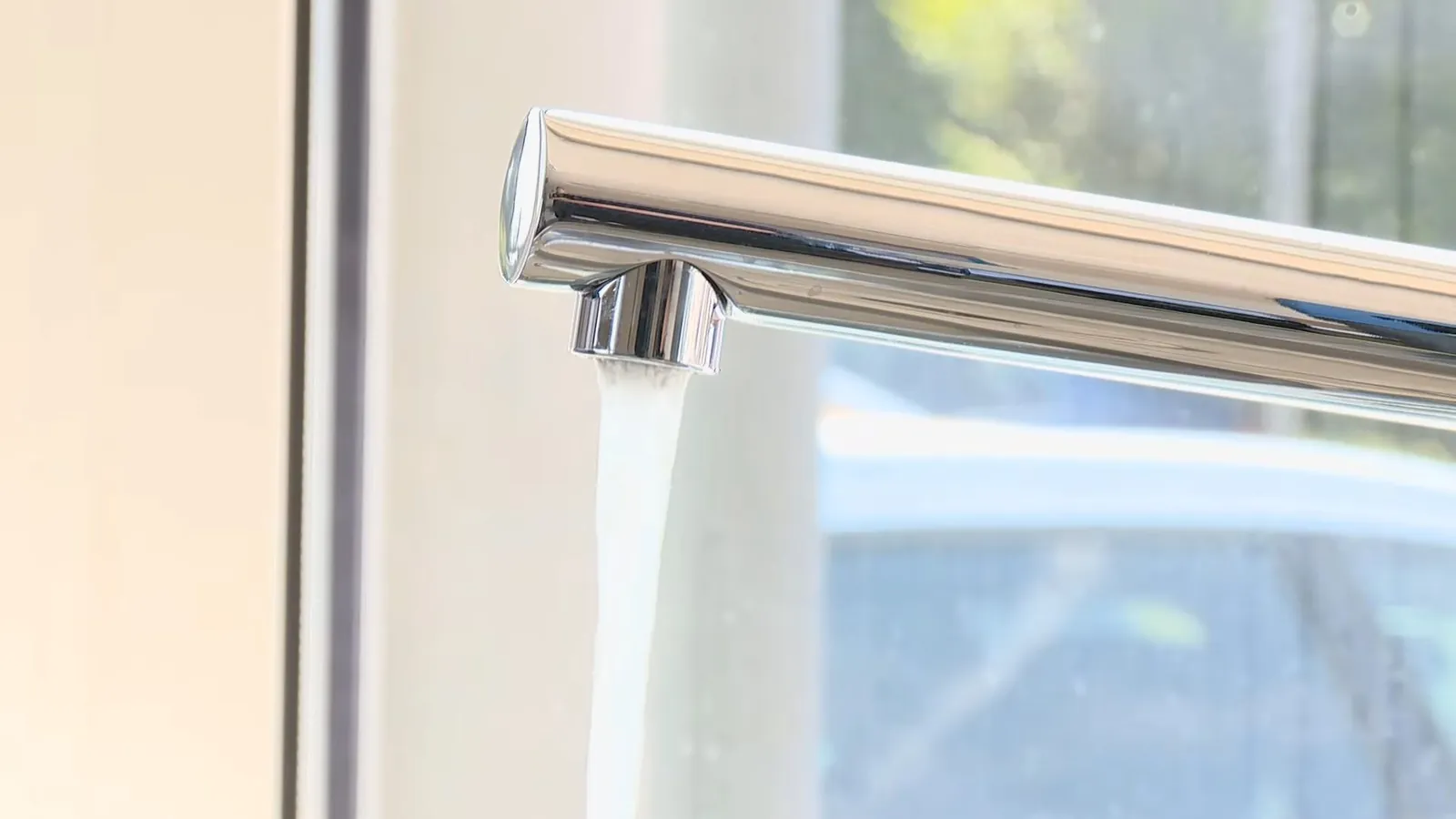 Chrome faucet spout with water running from it beside a window