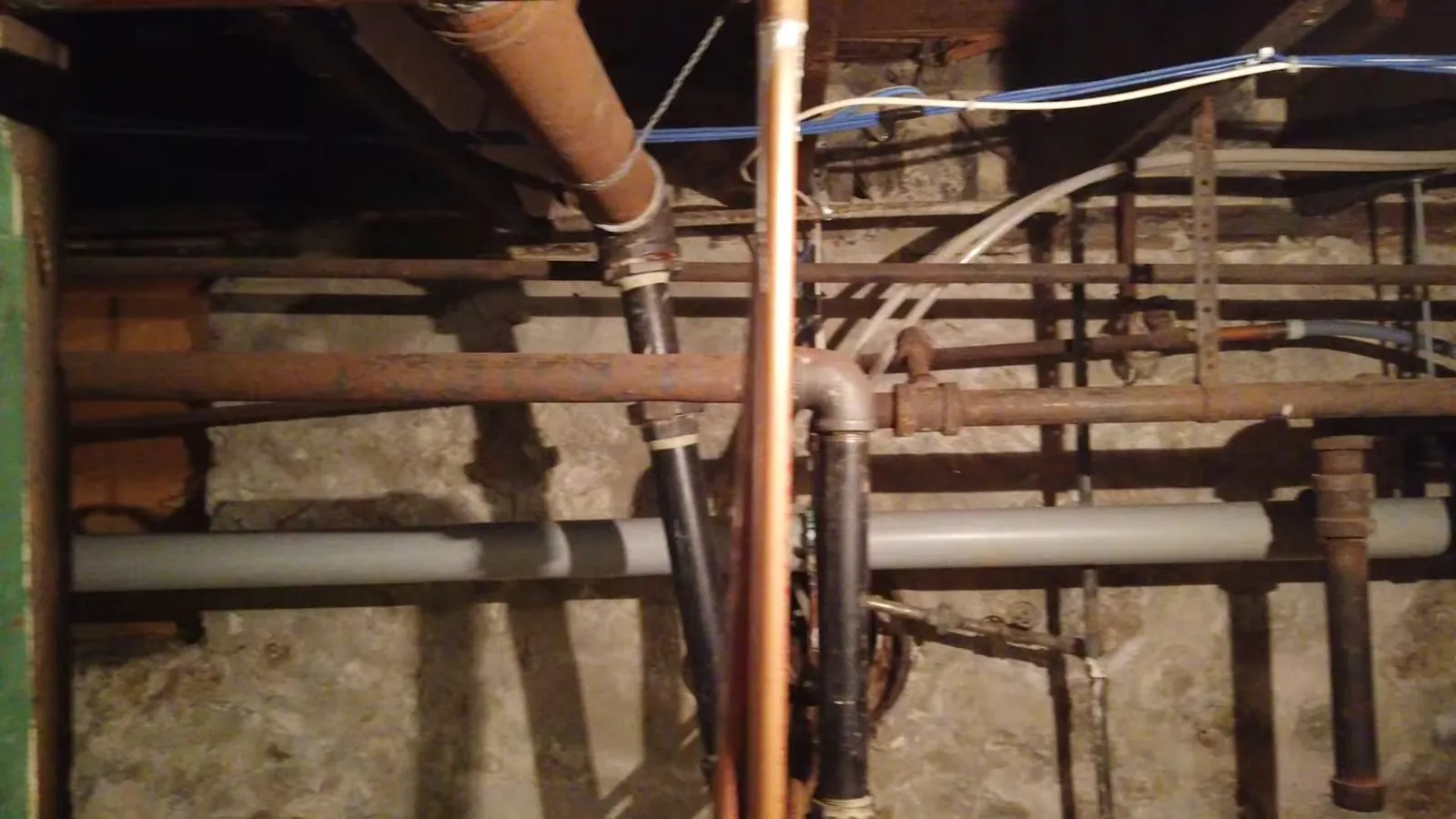 Rusty iron pipes and gray plastic pipes with a copper line running under old floor joists above a stone foundation