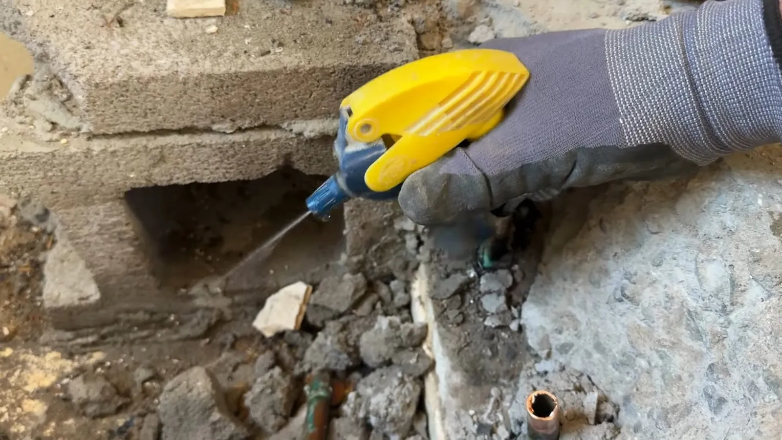 Gloved hand using a yellow tool on a small pipe in a broken section of concrete
