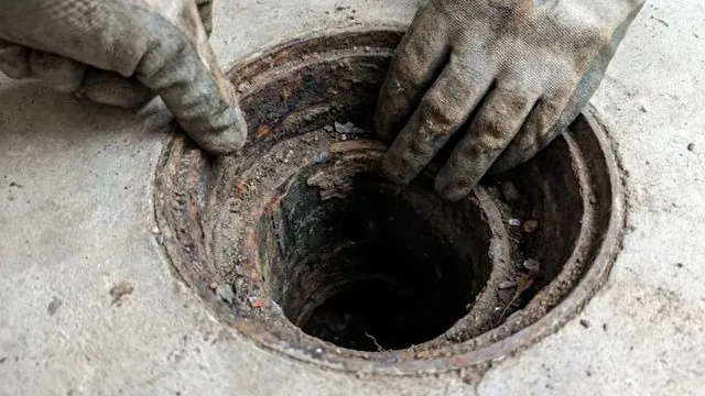 Two gloved hands lifting a rusty cover off a floor drain