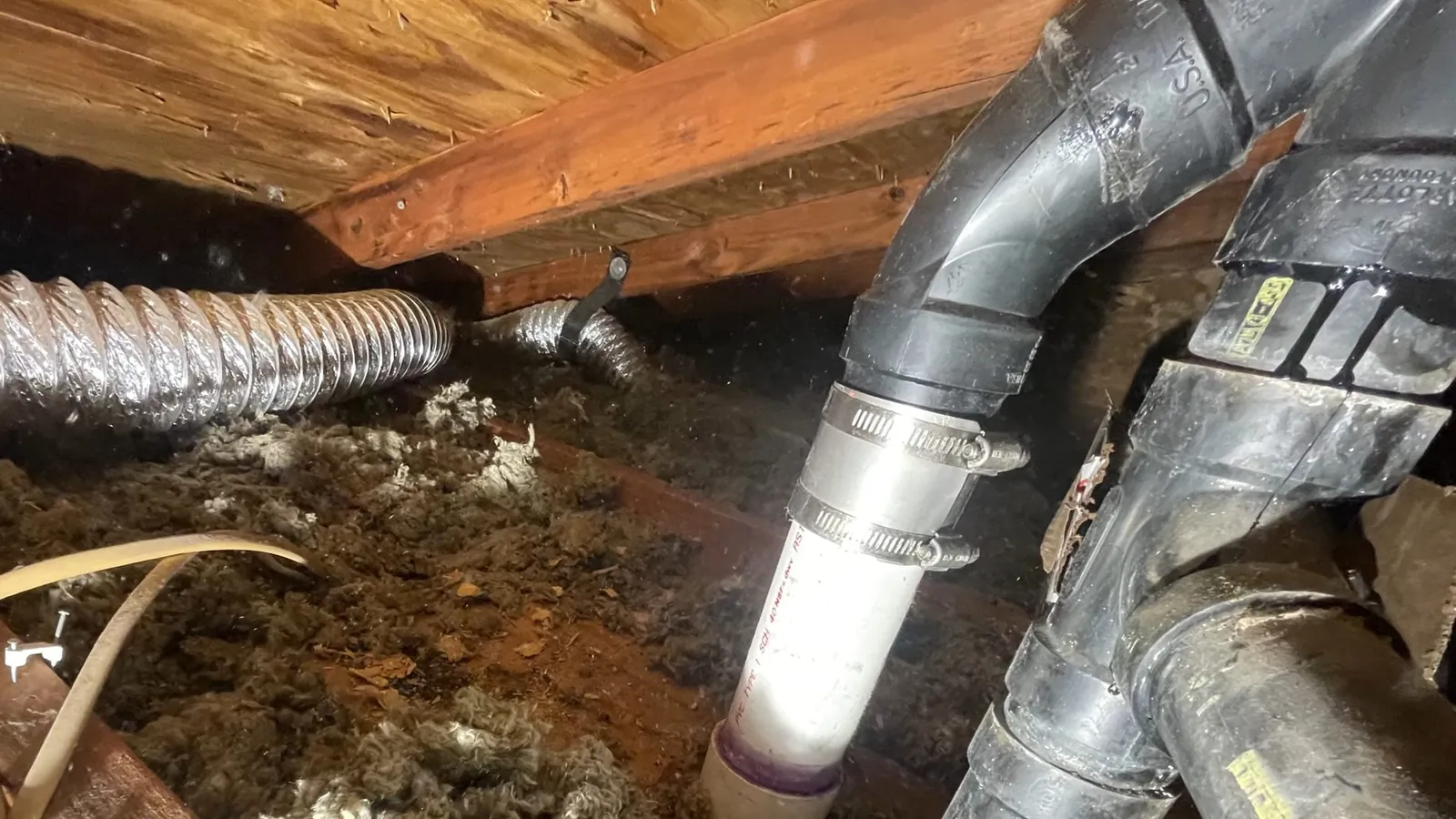 Black drain fittings and a white pipe among floor joists in a crawlspace, with silver flexible duct nearby