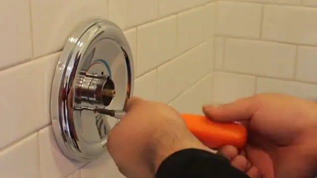 Hand turning an orange screwdriver on a chrome shower valve trim set in white tile