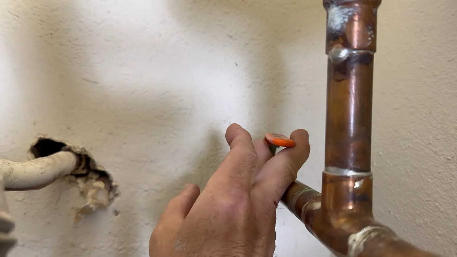 Hand holding an orange tool at a copper pipe that comes out of a drywall wall