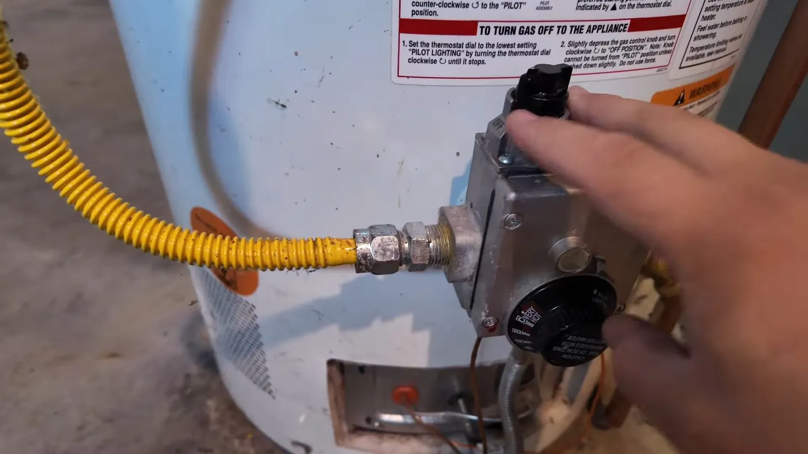 Hand on the gas control valve of a tank water heater beside a yellow flexible gas line