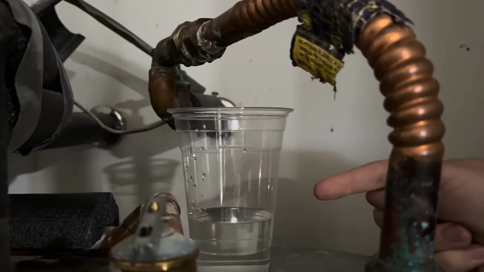 Hands holding a clear cup of water under a bent copper pipe with a valve above