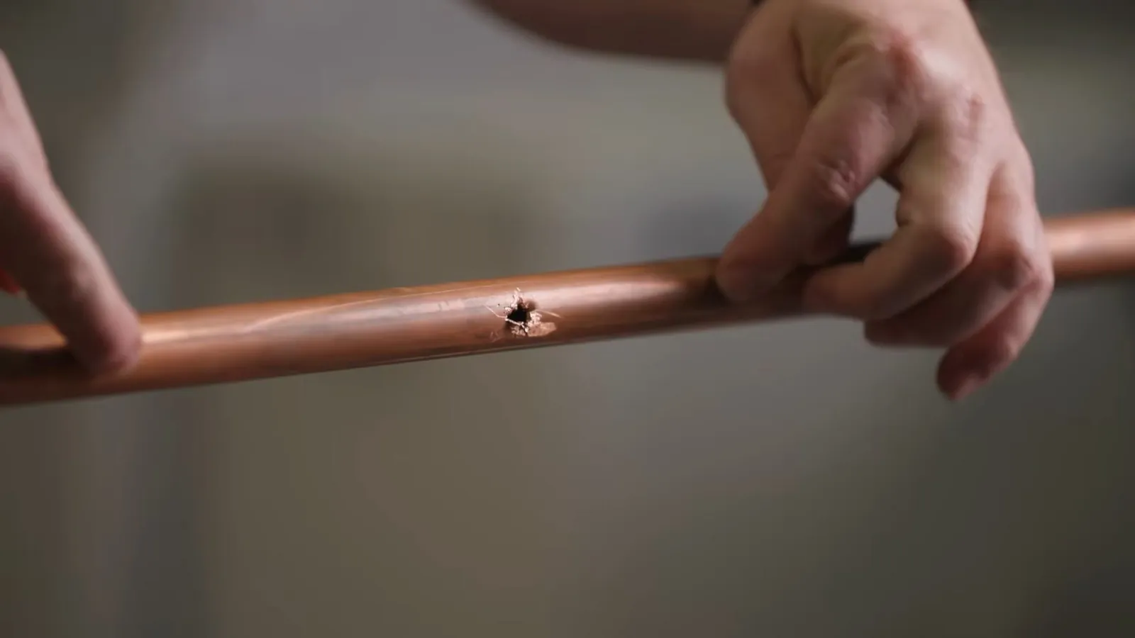 Hands holding a length of copper pipe with a small pinhole leak