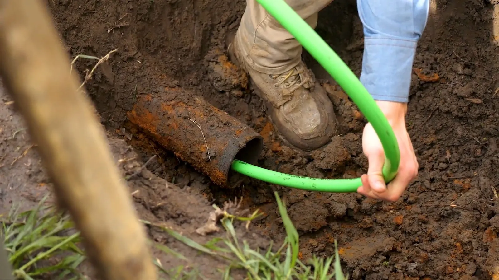 Man feeding a green hose into a rusty pipe opening in a dirt trench