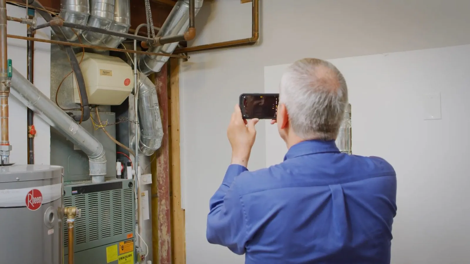 Man holding up a phone to photograph the pipes and tank in a water heater area