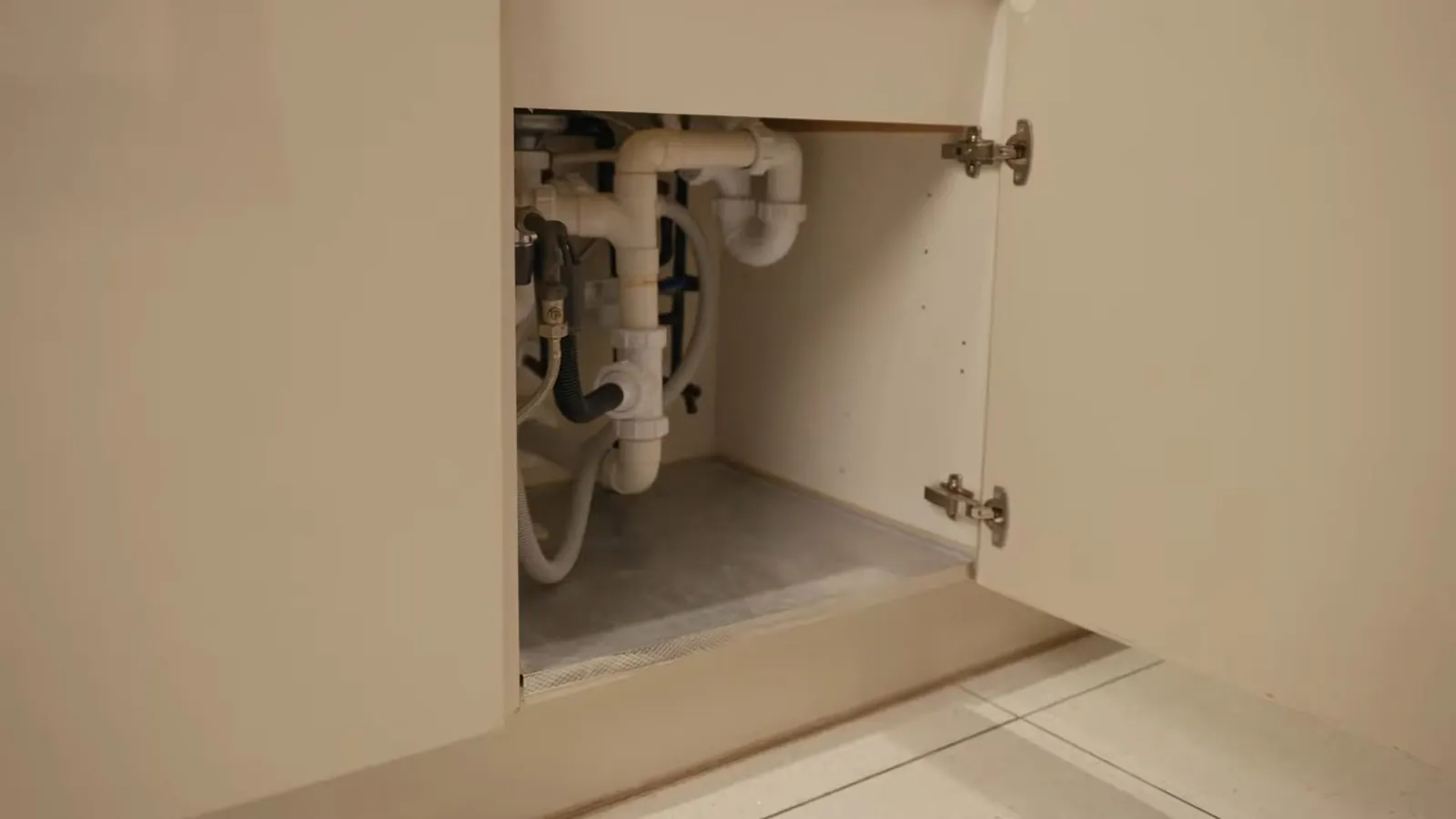 Open cabinet under a sink with white drain pipes and supply lines on the left side