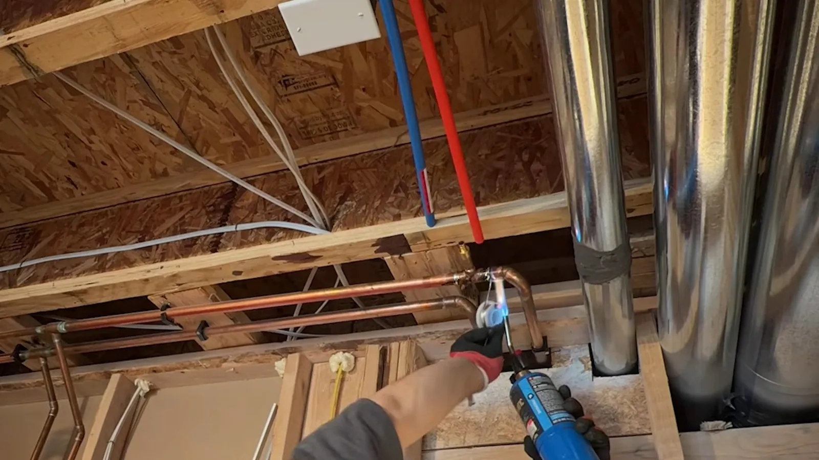 Open ceiling joists with red and white PEX lines and a hand holding a tool