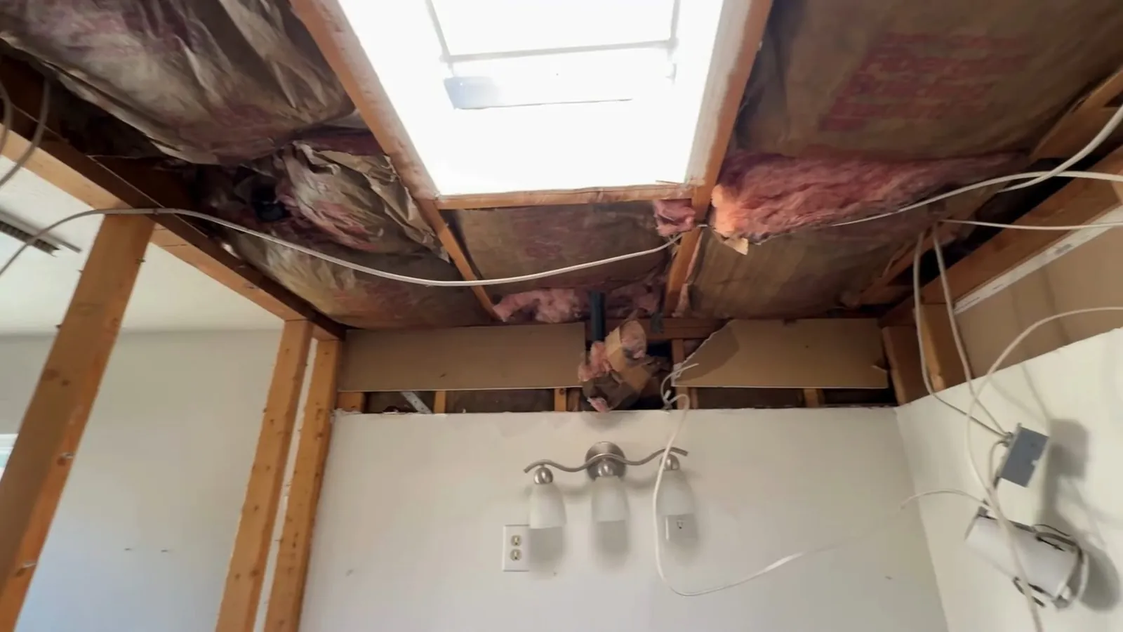 Opened bathroom ceiling showing pink insulation, wiring and framing around a skylight above a light fixture