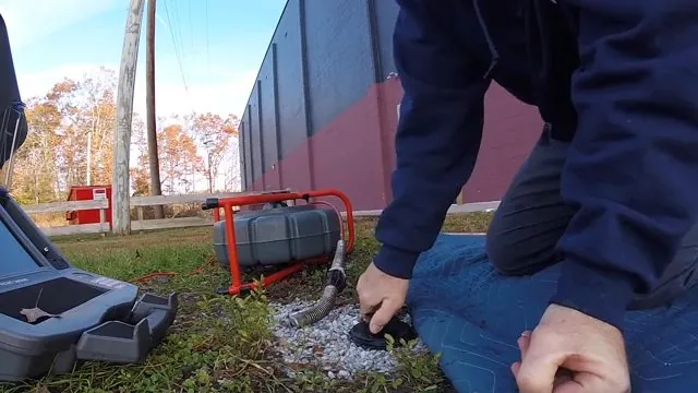 Two people kneeling in the grass beside a sewer camera reel at a cleanout near a fence