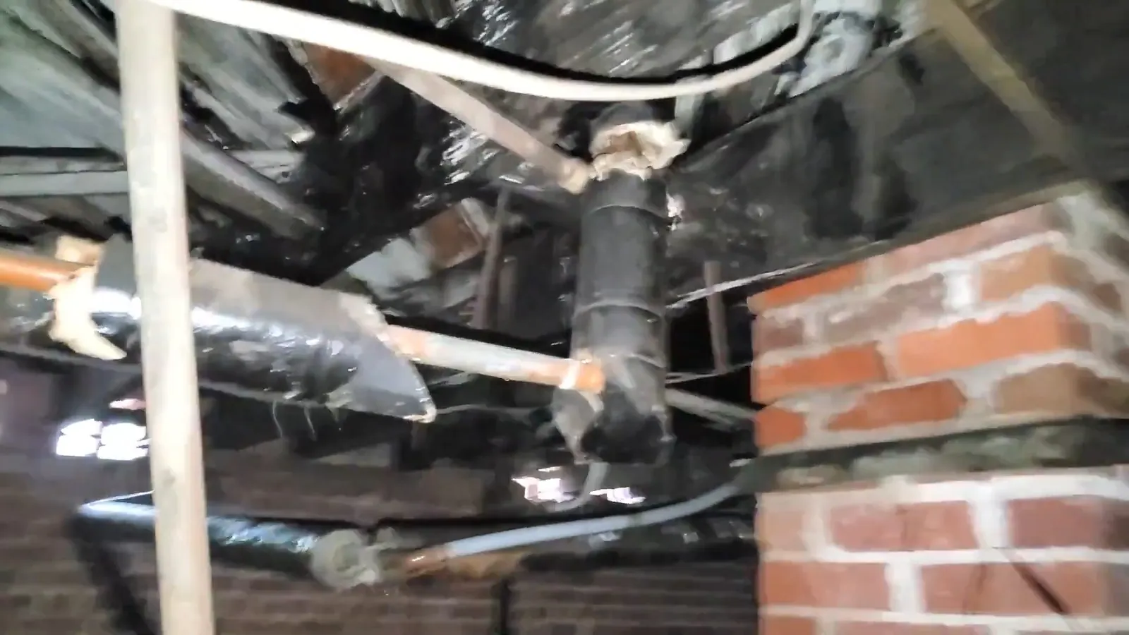 Black wrapped pipes and a vertical drain under a floor beside a brick pier