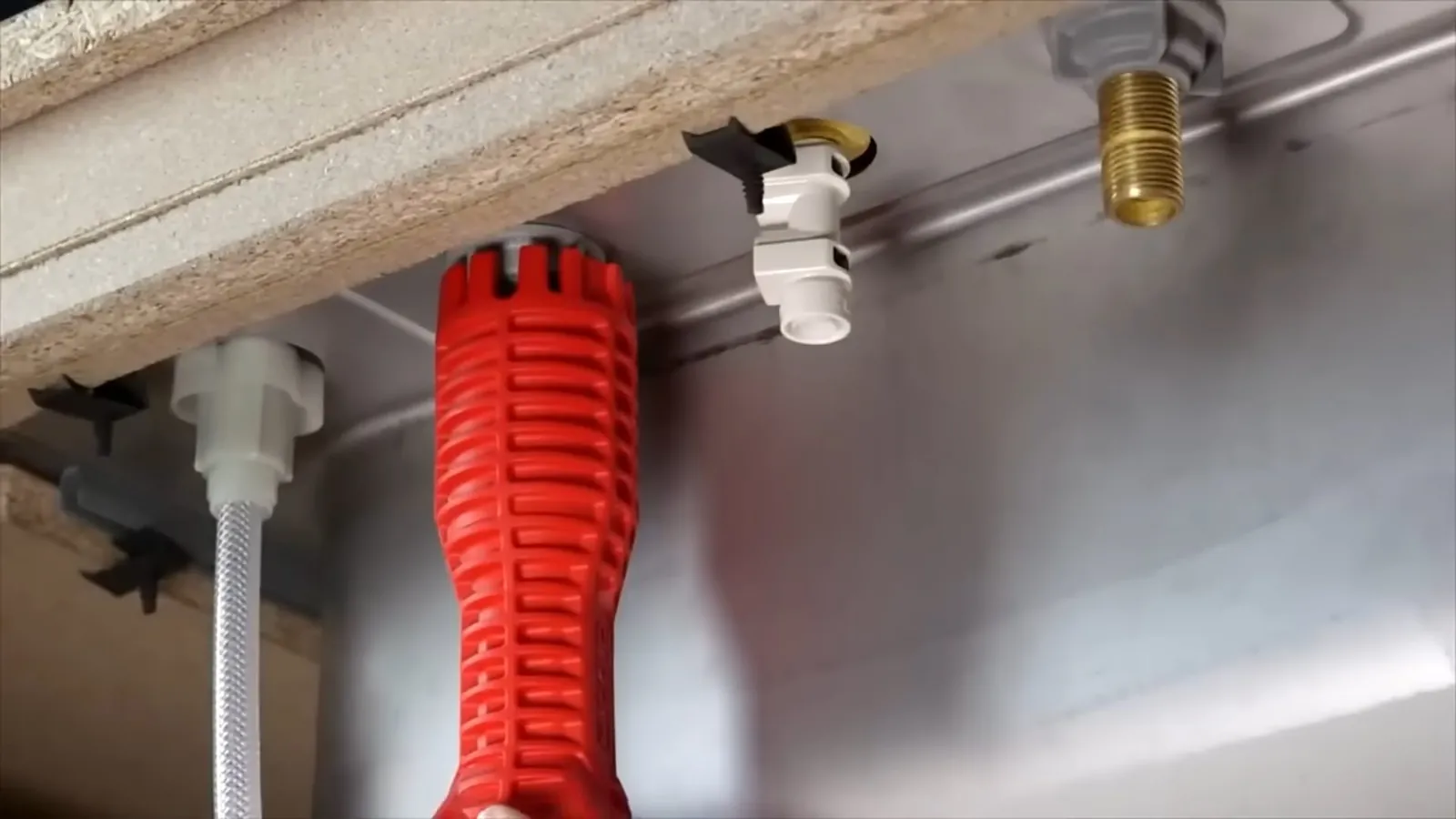 Red basin wrench resting under a sink beside supply lines and shutoff fittings