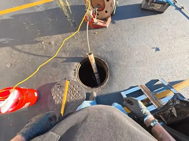 Sewer cleanout opening in pavement with a yellow cord running into it and gloved hands working beside it