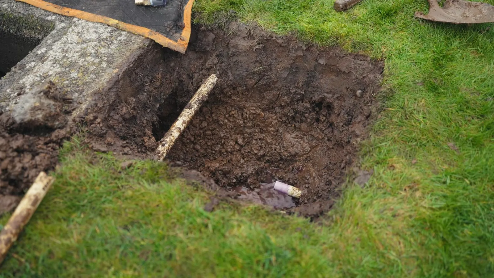Square hole dug in a grass lawn with a pale pipe visible at the bottom