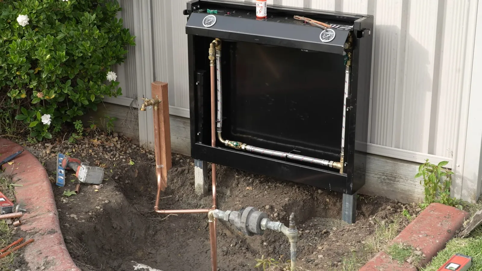 Tankless water heater mounted on an outside wall with copper water lines and a gas line below it