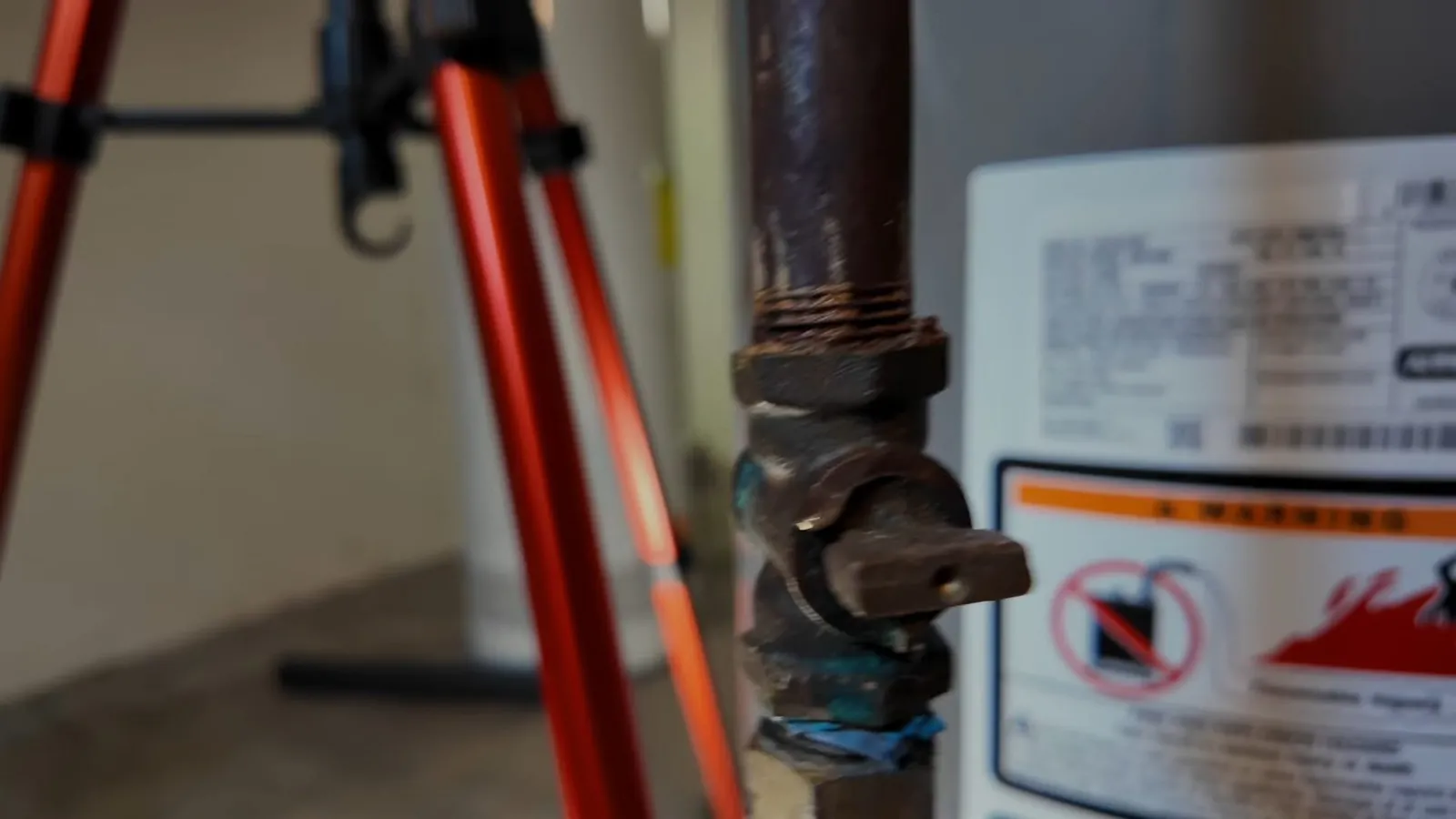 Old water heater with a rusty shutoff valve beside a red tripod
