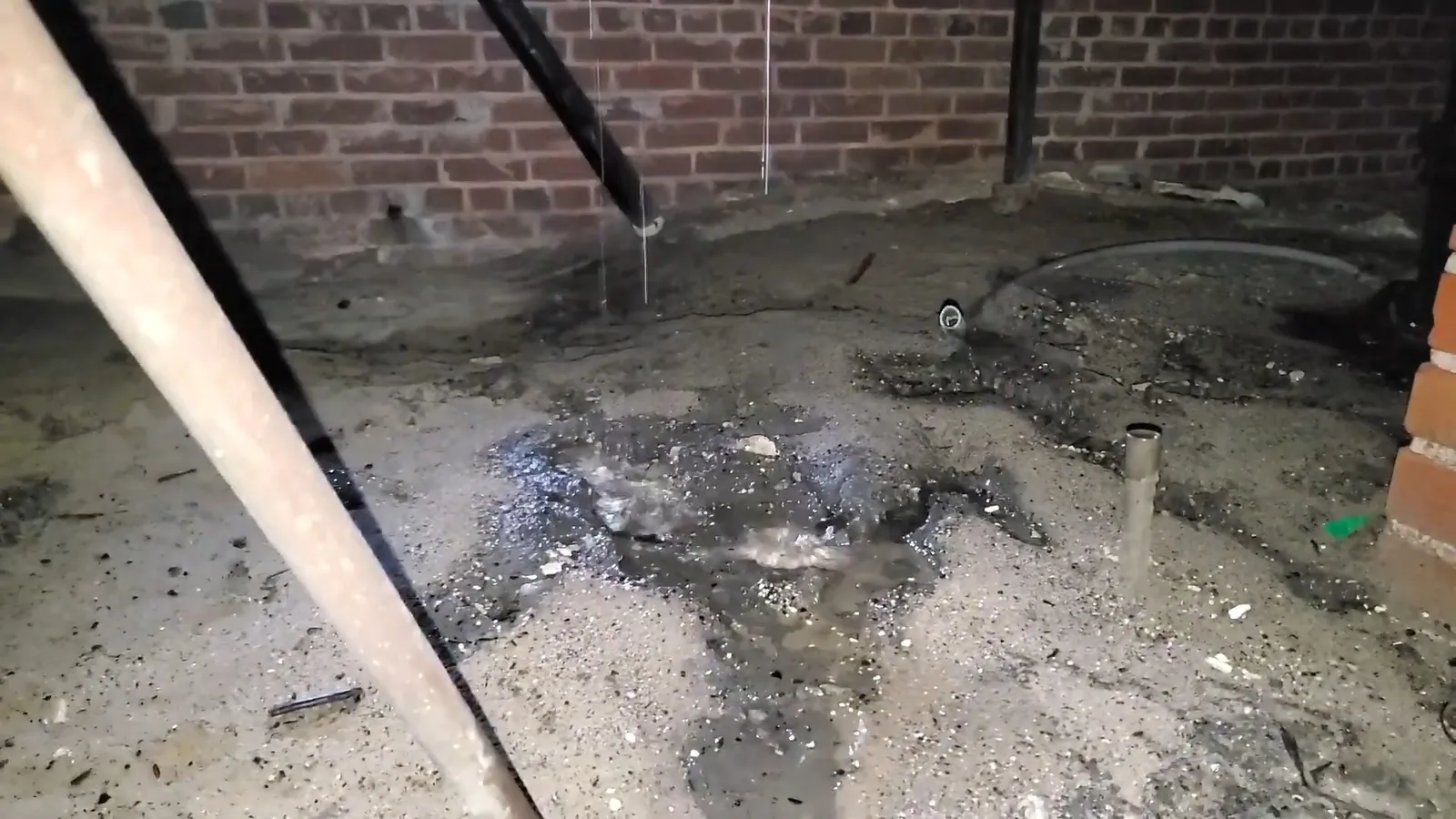 Wet concrete floor with a pipe stub and debris beside a brick wall