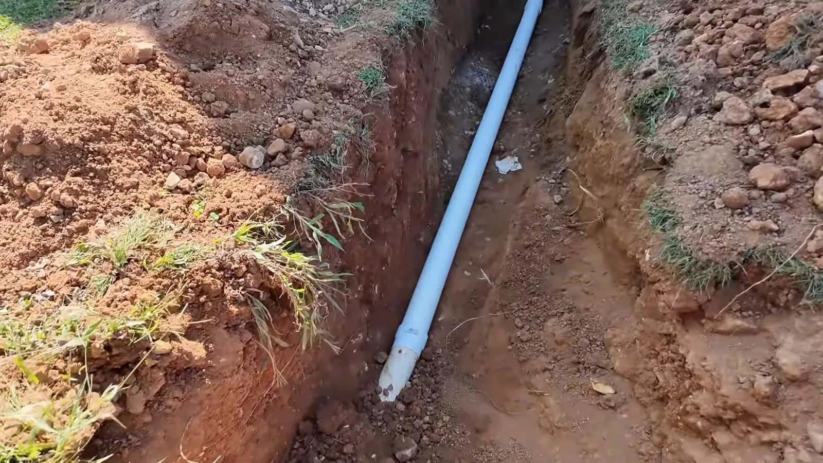 Narrow dirt trench with a white pipe lying at an angle along the bottom and grass along the edges