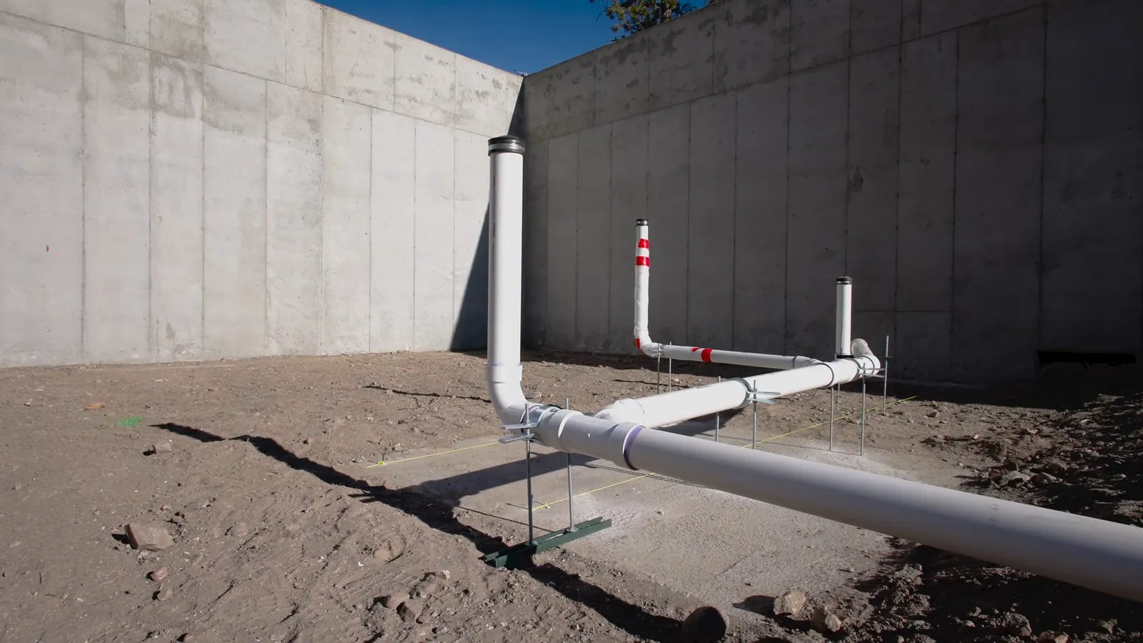 White PVC drain pipes with vertical cleanout risers standing in a dirt lot beside a tall concrete wall