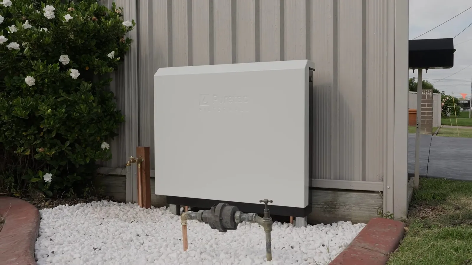 White utility box and pipes on a gravel bed beside a corrugated metal fence with white flowers
