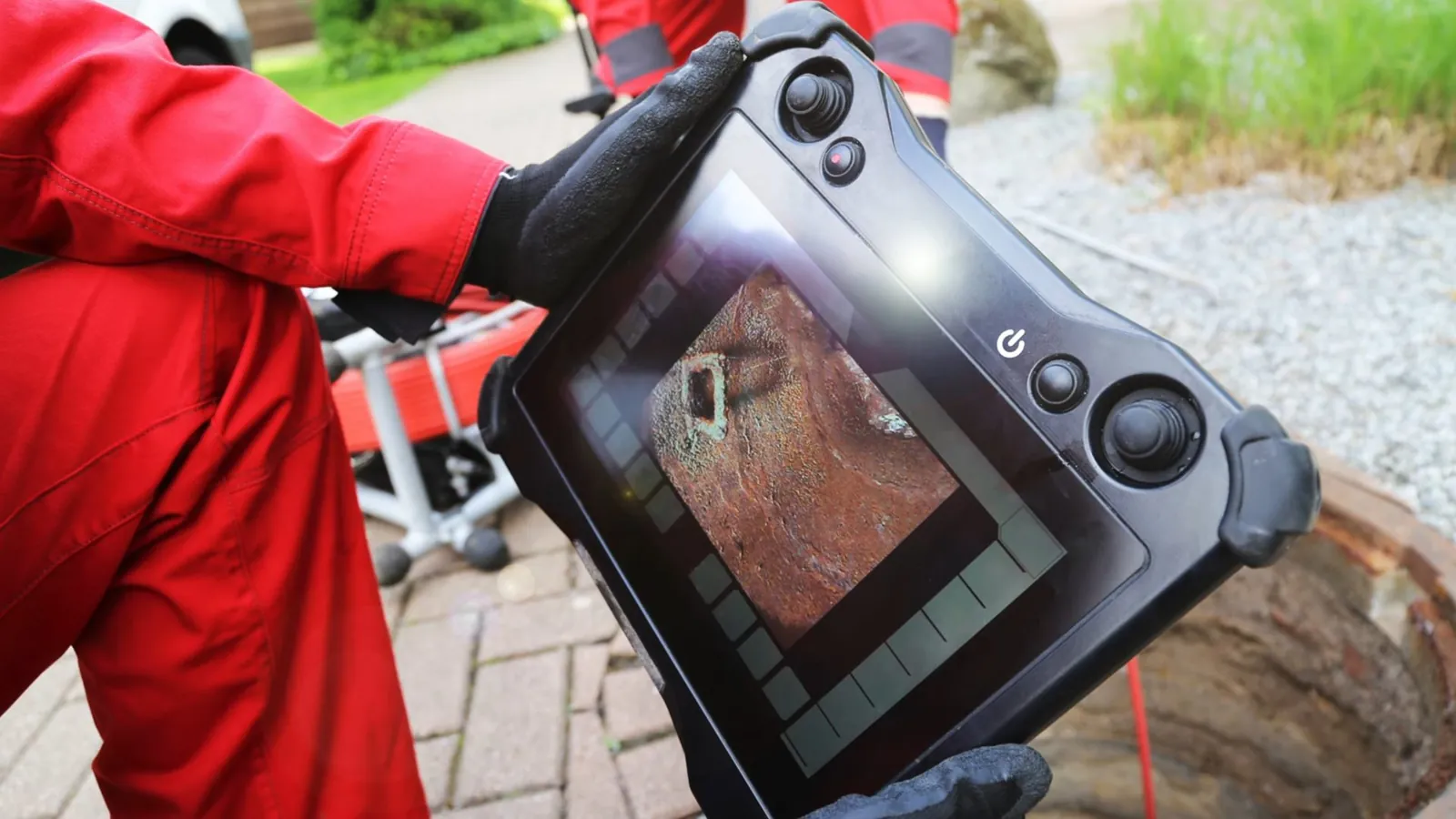 Worker in red holding a handheld sewer camera monitor beside a brick paved area